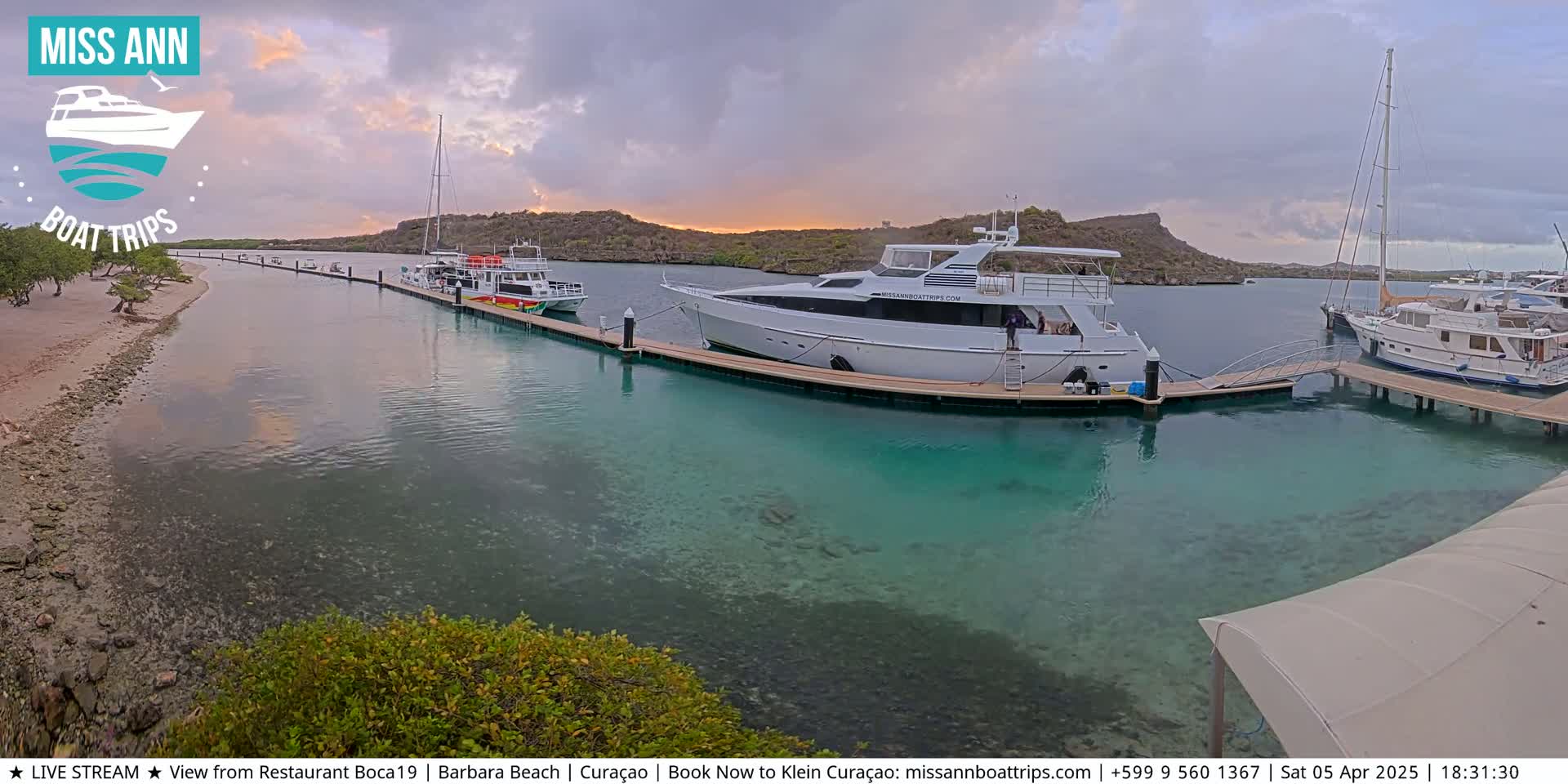 Several boats are docked at a marina with clear, turquoise water under an overcast sky.