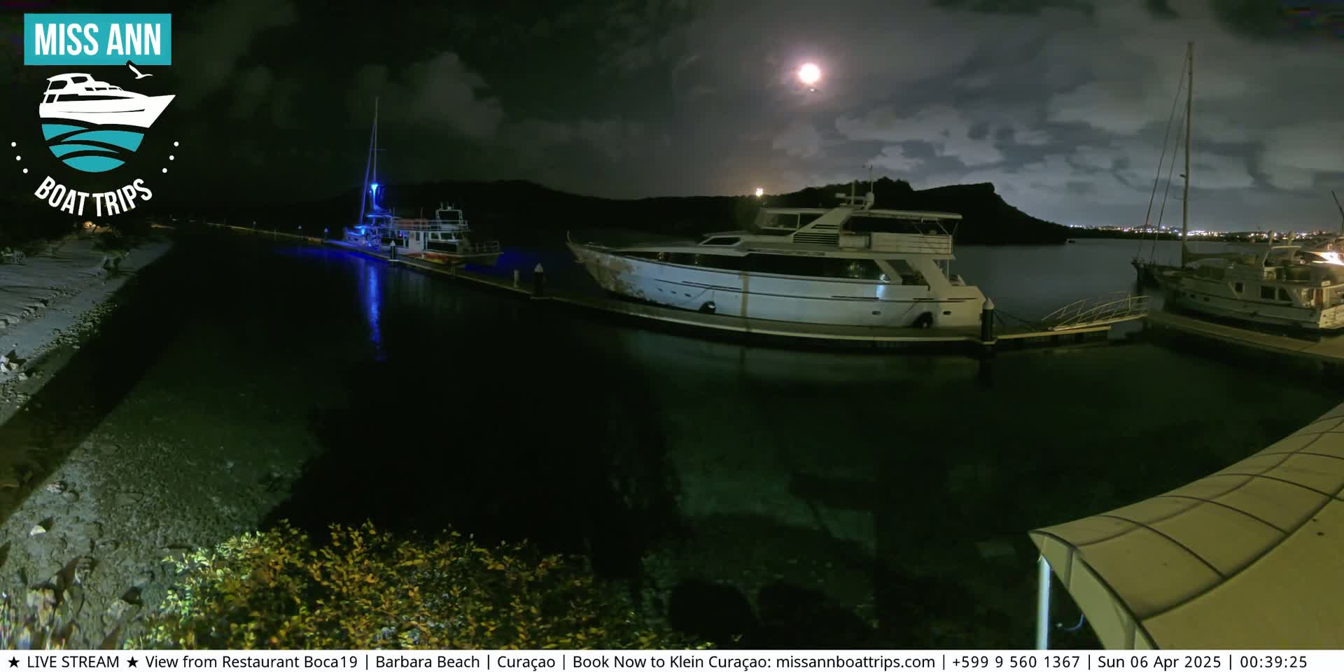 Several boats are moored at a marina at night under a partly cloudy sky, illuminated by a bright moon.