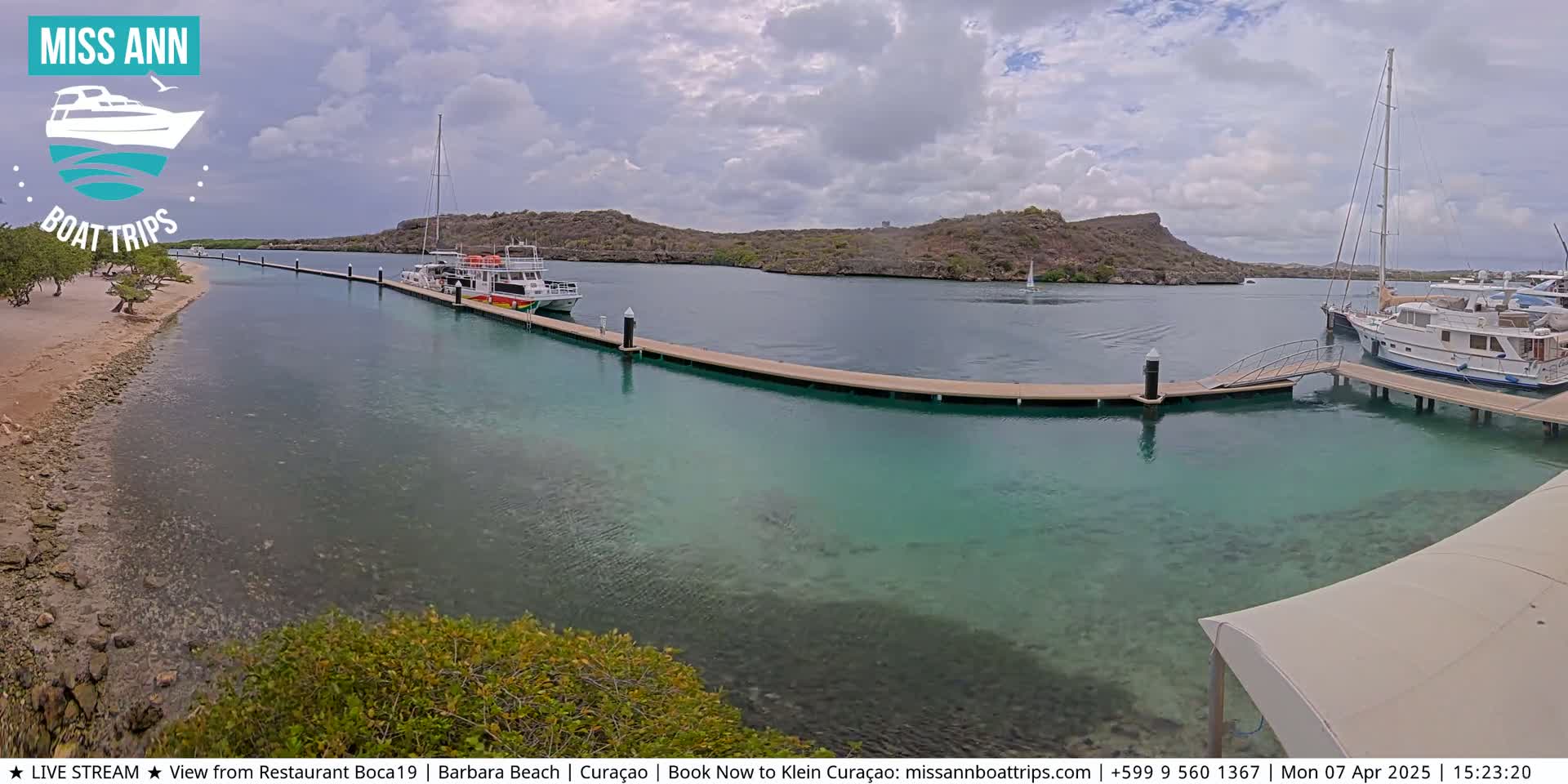 A calm, partly cloudy day shows a marina with several boats docked at piers, a sandy beach, and clear, shallow turquoise water surrounding rocky land.