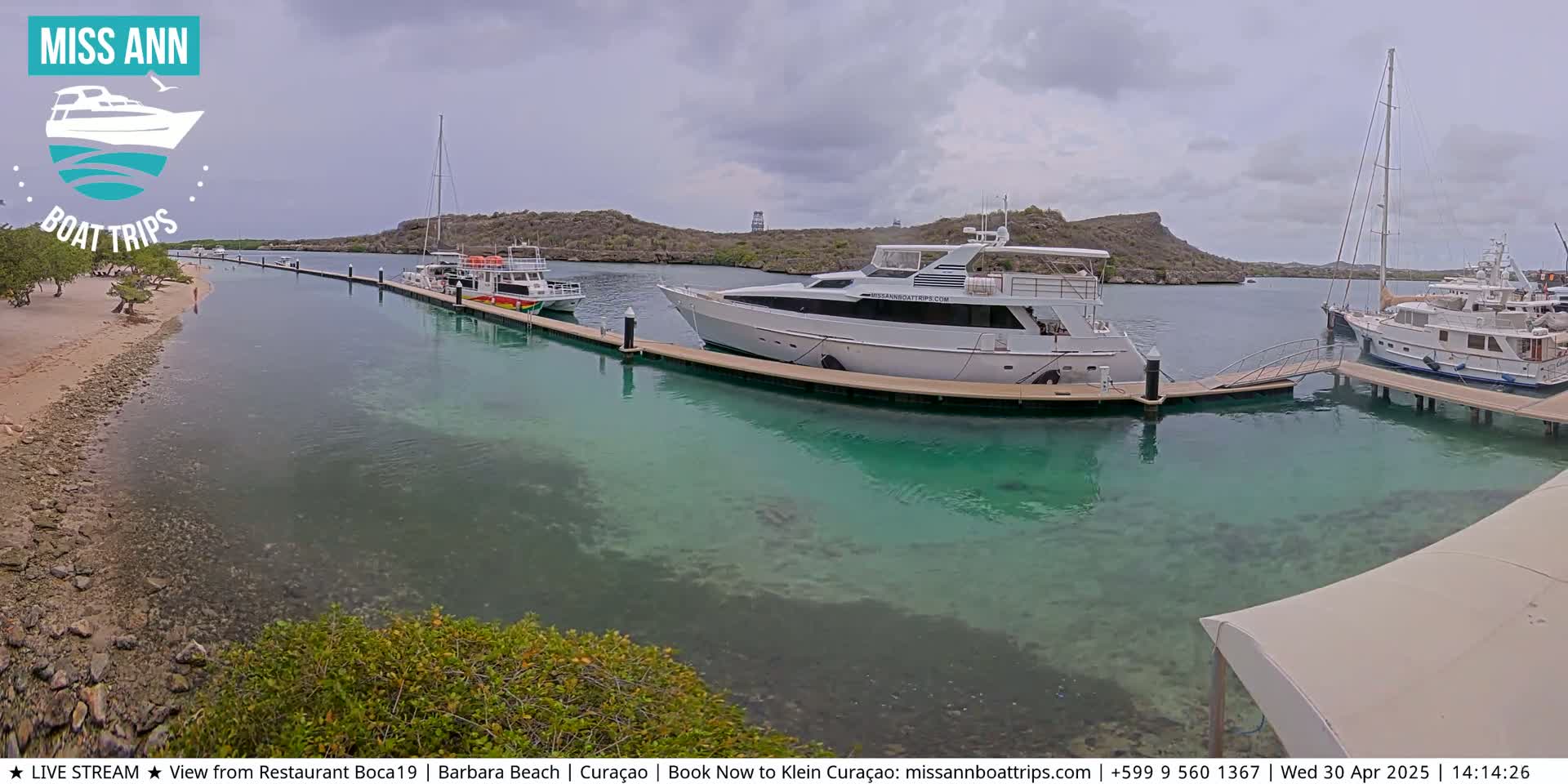 Several boats are docked at a marina on a partly cloudy day, with clear, shallow turquoise water visible around the docks and a sandy beach in the background.