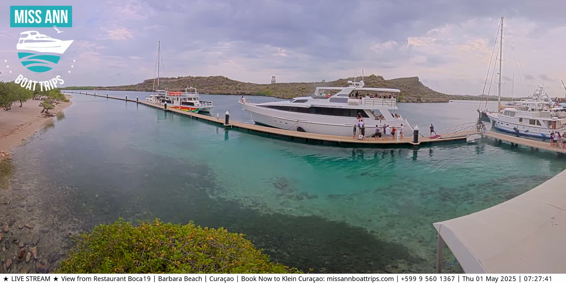 Several boats are docked at a marina with clear, shallow turquoise water, under a partly cloudy sky.