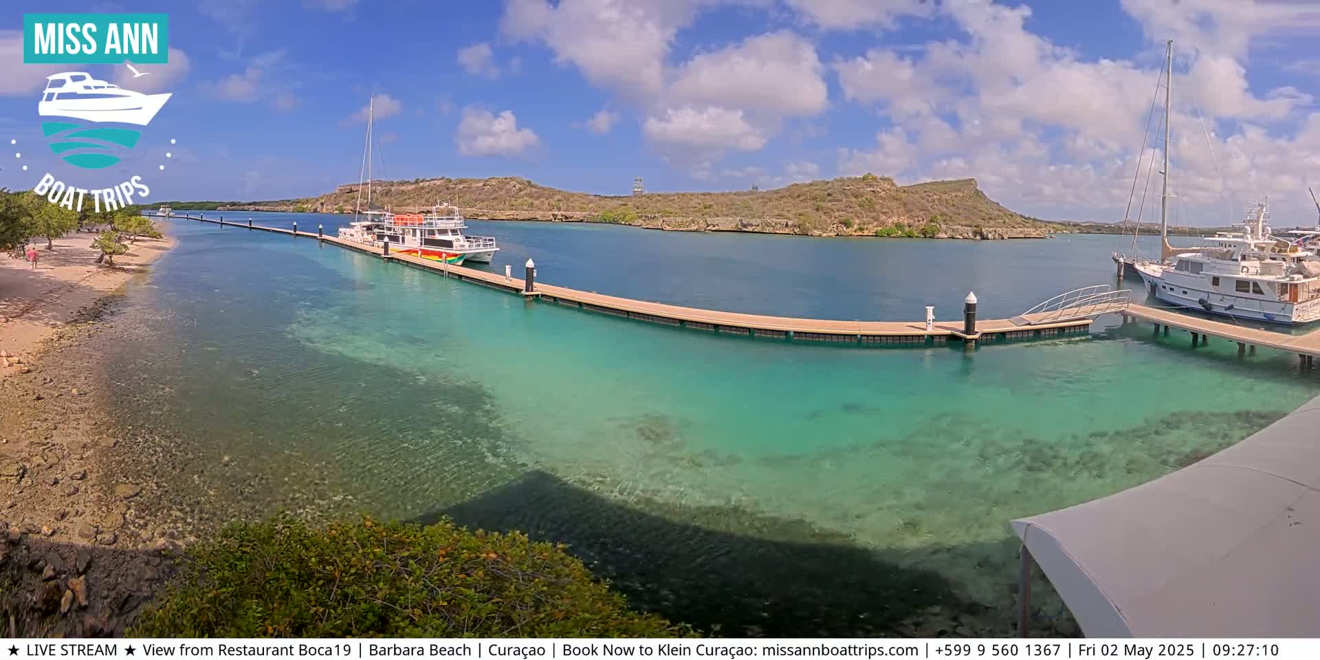 A sunny day reveals a calm, turquoise bay with several boats docked at a pier near a sandy beach and a small island.
