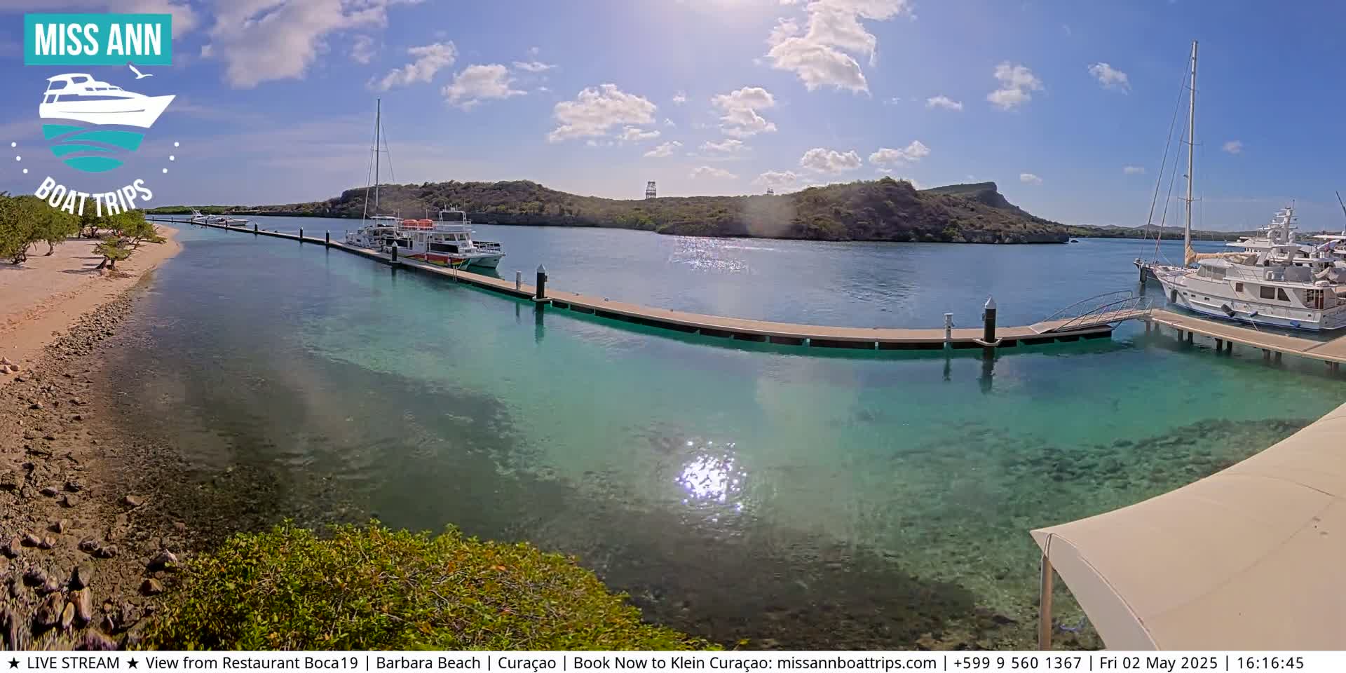 Several boats are moored at a dock in clear, shallow turquoise water, under a sunny sky with a few clouds.