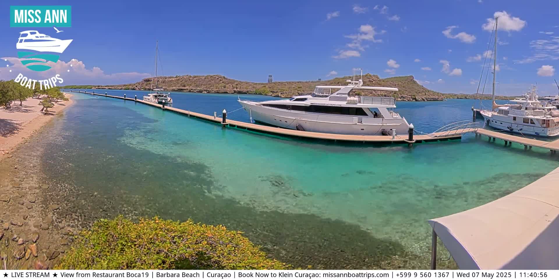 Several yachts are docked at a marina with clear, shallow turquoise water, under a sunny sky.