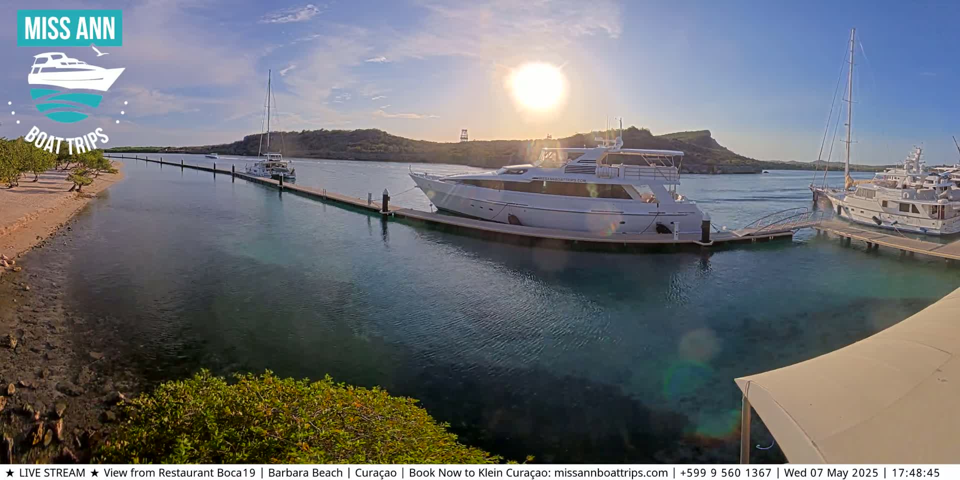 Several boats are docked at a marina on a sunny day, with calm, clear water and hills visible in the background.