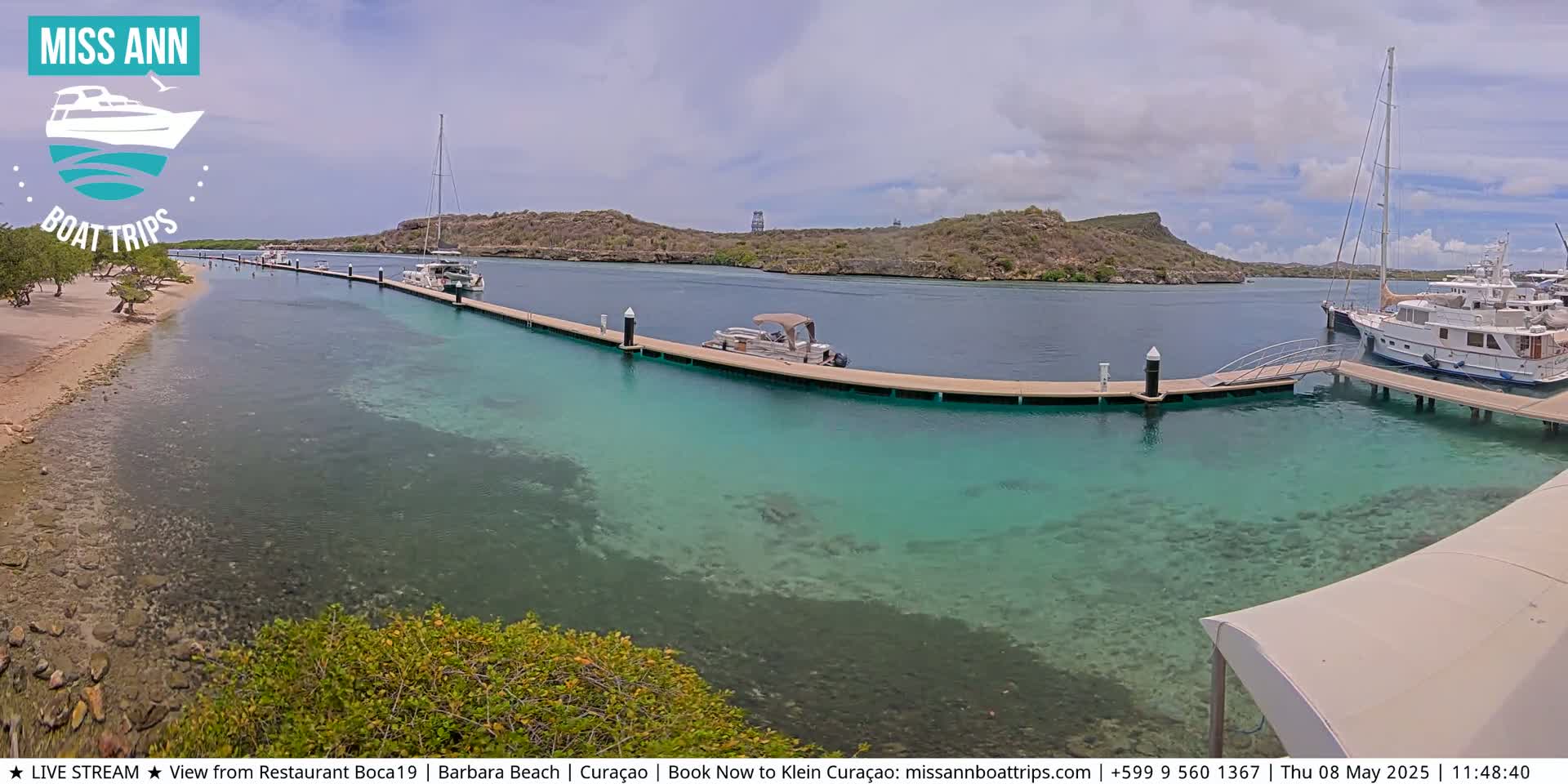 A calm, sunny day shows a marina with boats docked at a long pier extending into clear, shallow turquoise water, surrounded by a sandy beach and low-lying hills.
