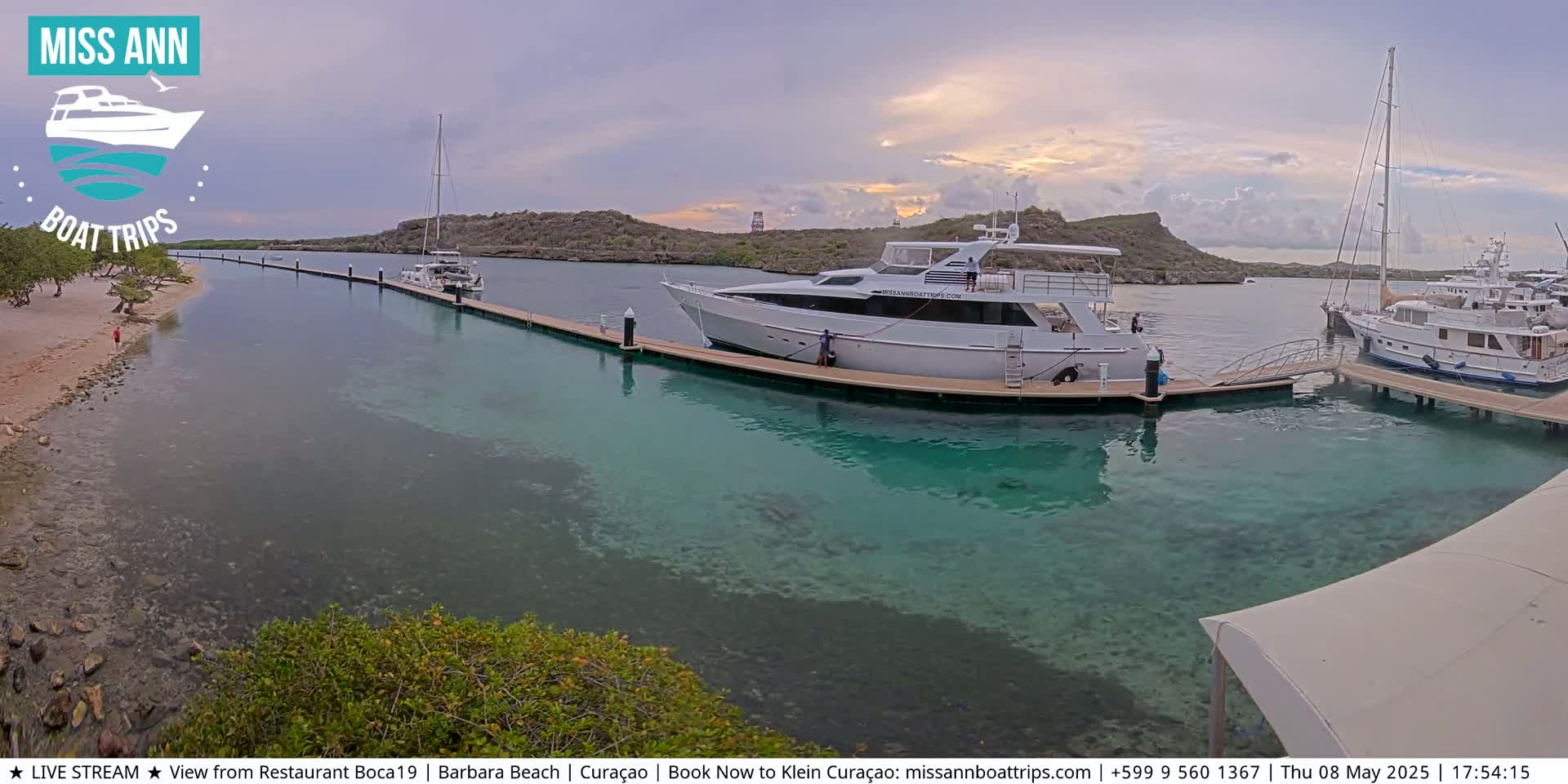 Several boats are docked at a marina with clear, shallow turquoise water, under a partly cloudy sky.