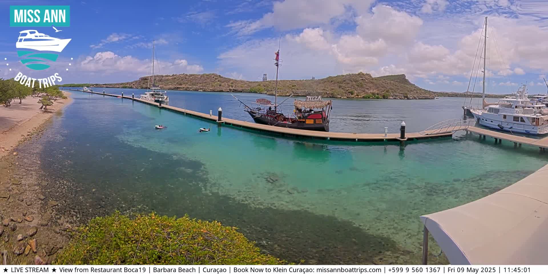 A pirate ship is docked at a marina on a sunny day with clear, turquoise water, surrounded by other boats and a sandy shoreline.