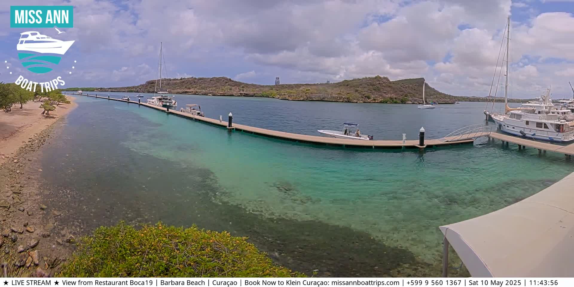 A calm, clear turquoise body of water shows a marina with several boats docked under a partly cloudy sky.