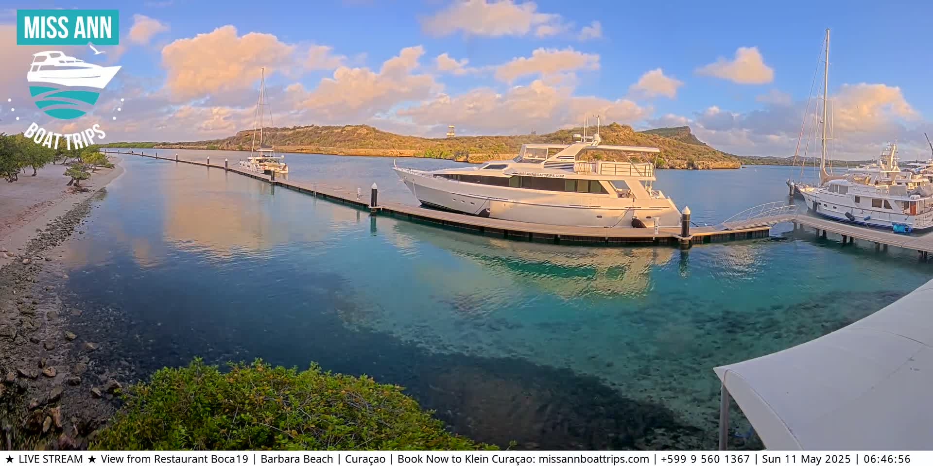 A large yacht is docked at a marina, with other boats and calm, clear turquoise water under a partly cloudy, sunset sky.