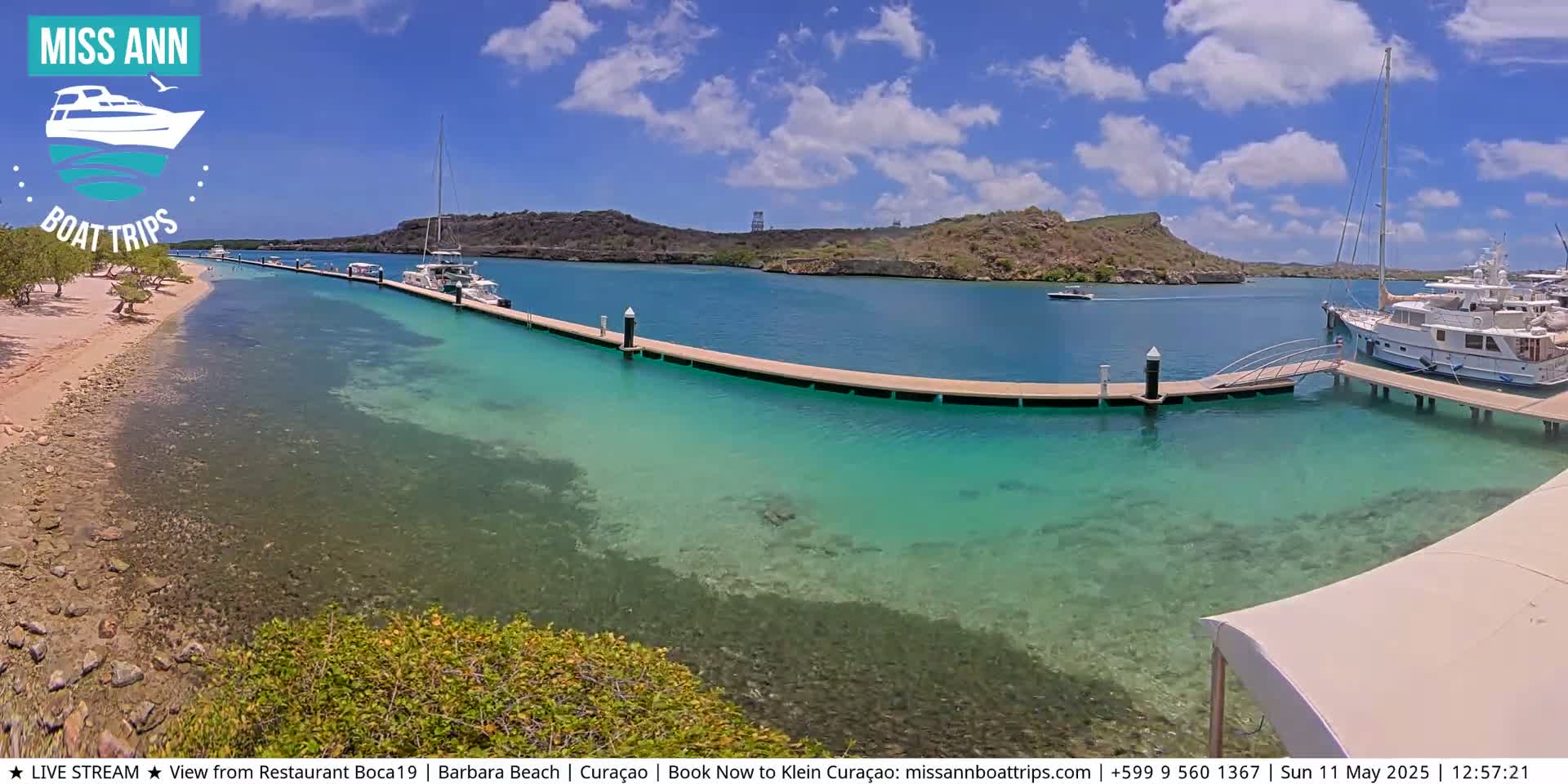 A long dock extends into clear, turquoise water, lined with boats near a sandy shore and rocky hills under a sunny sky.