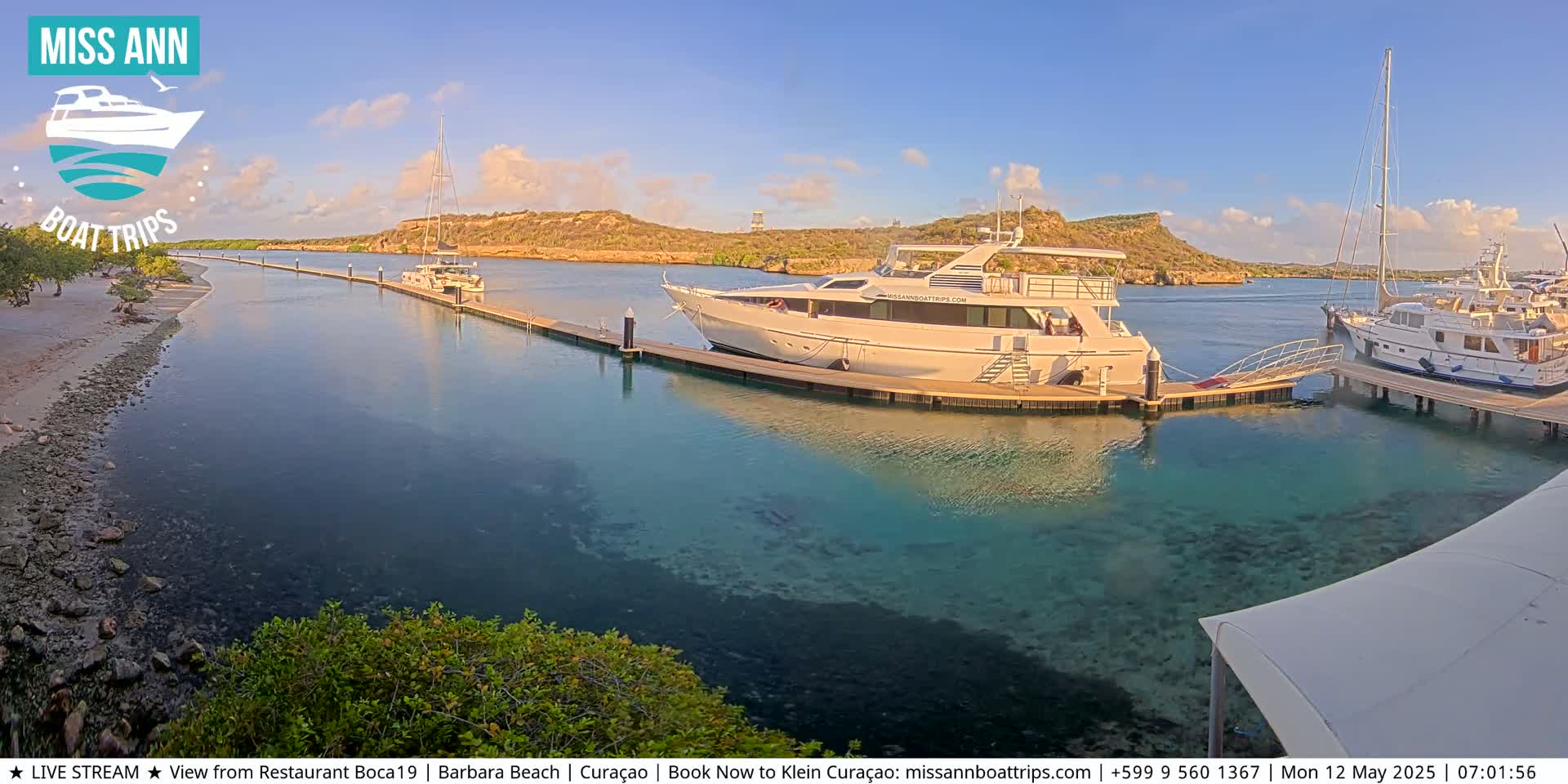 Several boats are docked at a marina on clear, shallow turquoise water, with a sandy shore and hills in the background under a mostly sunny sky.