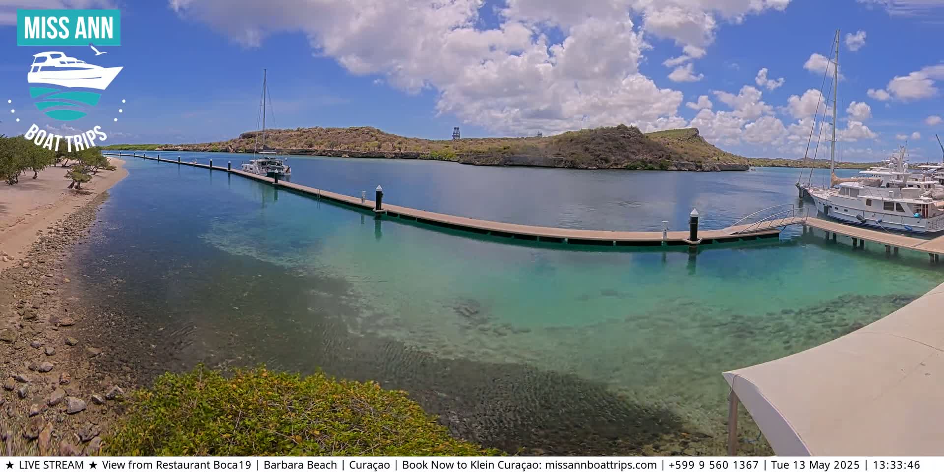 A partly cloudy day shows a calm, clear turquoise body of water with several boats docked at a long pier next to a sandy shore and low-lying vegetation.