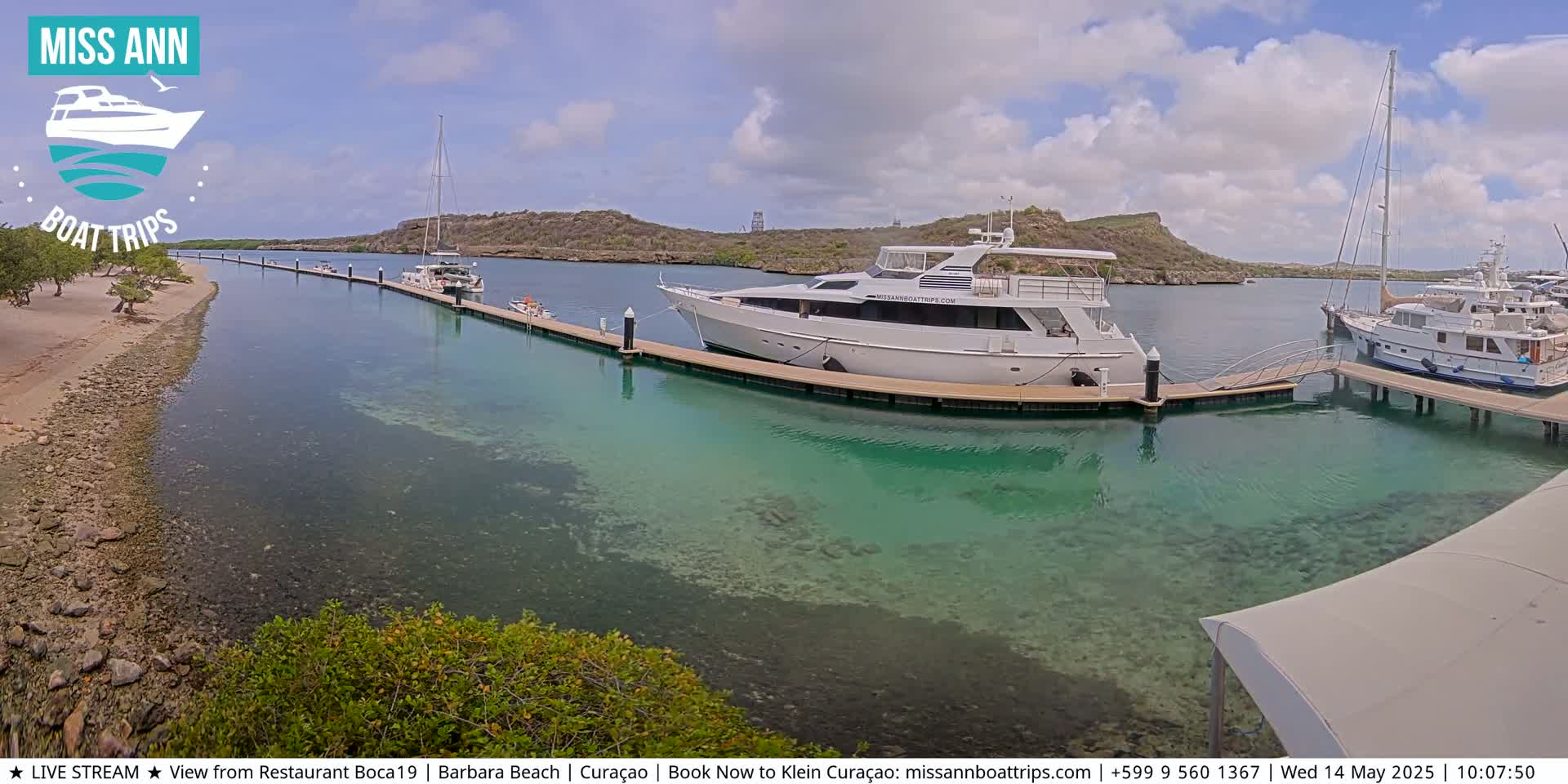 A marina with several boats docked, clear shallow turquoise water, and a sandy shore with vegetation in the background under a partly cloudy sky.