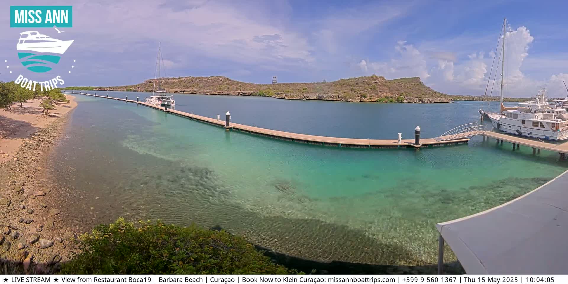 A clear, sunny day shows a calm, turquoise body of water with several boats docked at a long pier near a sandy shore and rocky hillside.