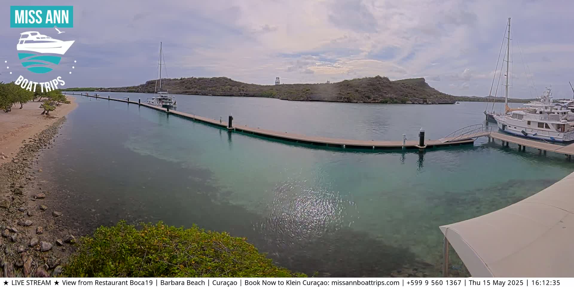 A calm, partly cloudy day shows a marina with several boats docked at a long pier, extending from a sandy beach to a small, rocky island.