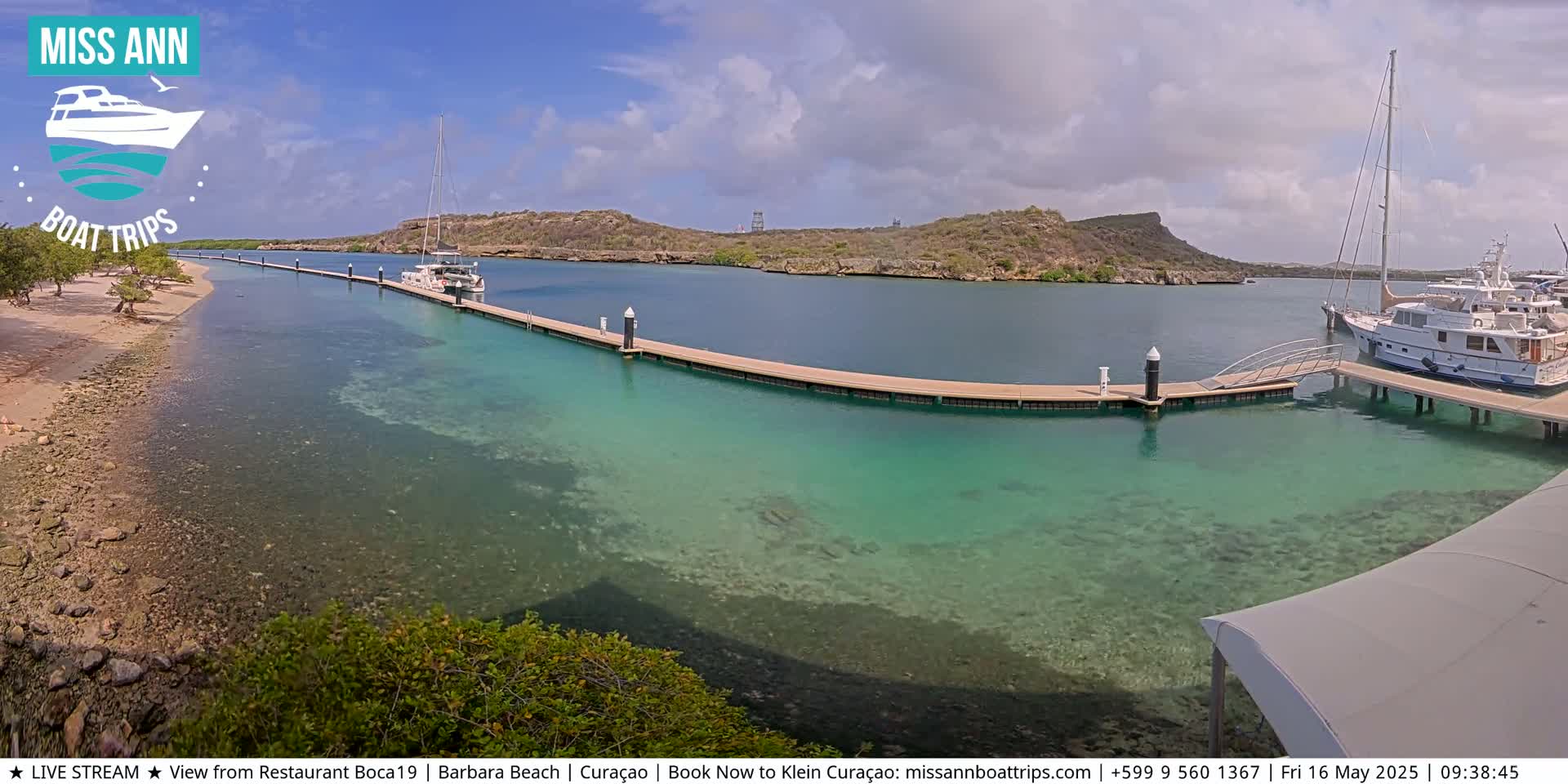 A marina with clear turquoise water, boats docked at piers, and a sandy shoreline under partly cloudy skies.