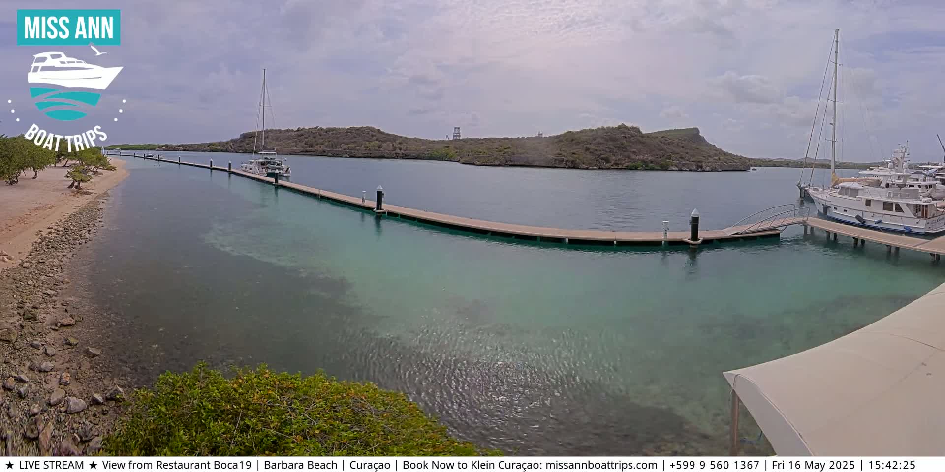 A calm, partly cloudy day shows a marina with several boats docked at a long pier extending into clear, shallow turquoise water beside a sandy beach.