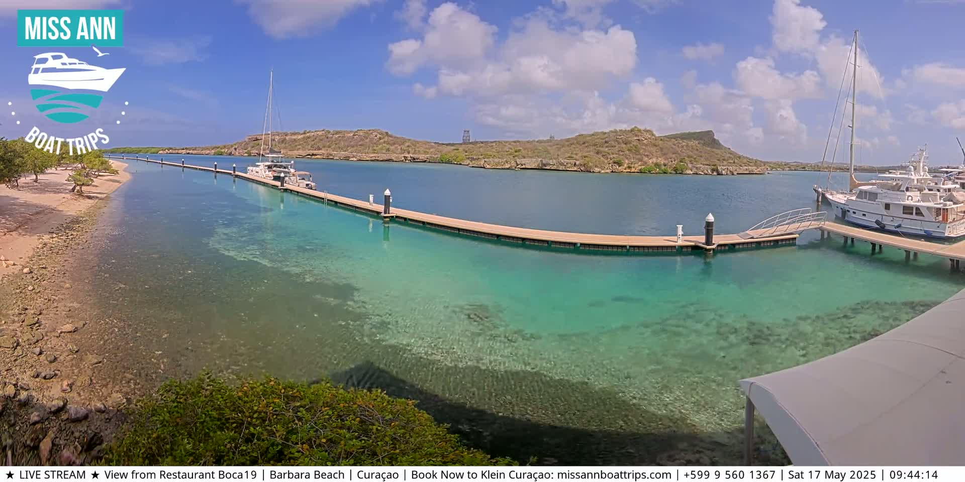 A sunny day reveals clear, shallow turquoise water surrounding a marina with several boats docked at a long pier, near a sandy beach and rocky shoreline backed by low hills.