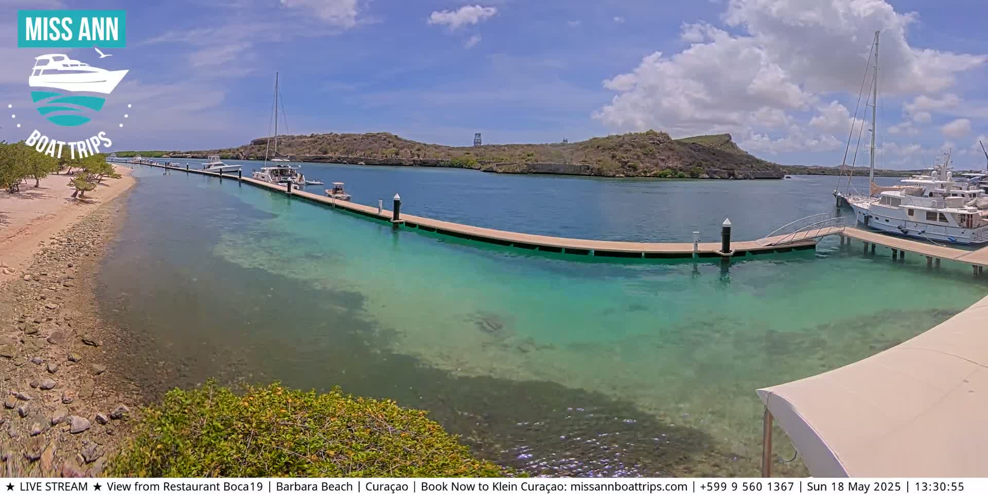 A sunny day shows a calm, clear turquoise body of water with several boats docked at a long pier, next to a sandy beach and low-lying hills.