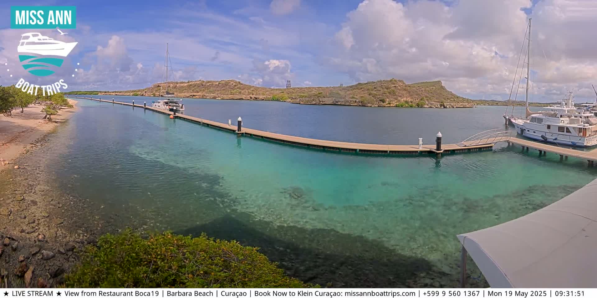 A partly cloudy day shows a calm, shallow turquoise bay with a long dock extending from a sandy shore, where several boats are moored.