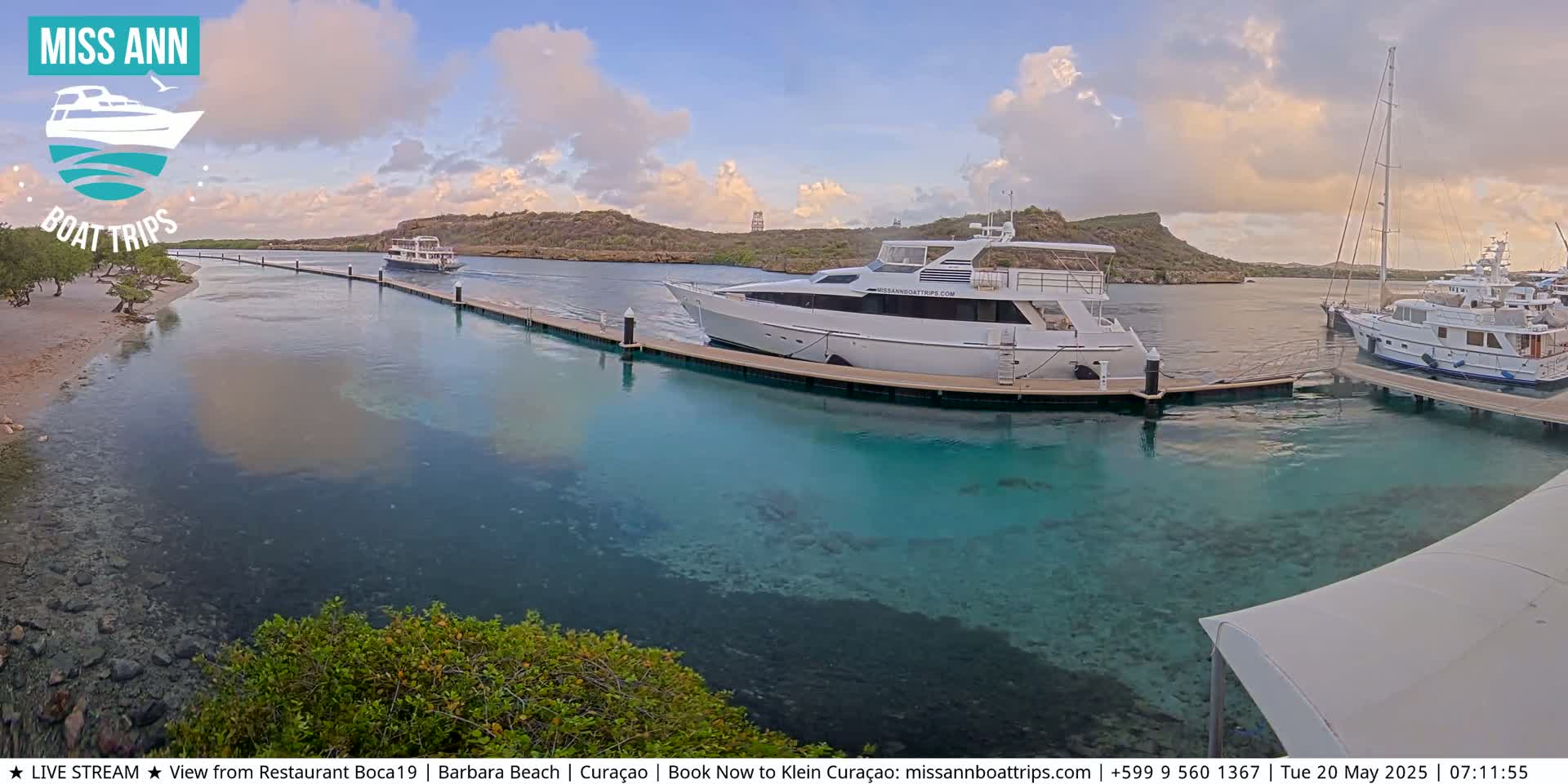 Several boats are docked at a marina with clear, shallow turquoise water, under a partly cloudy sky.