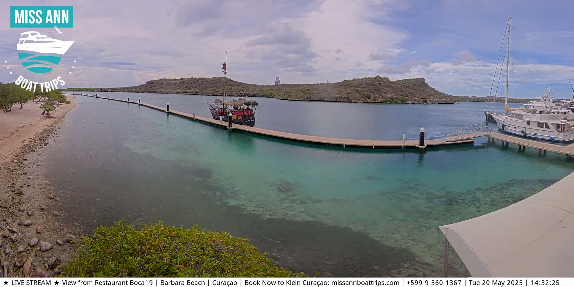 A partly cloudy day shows a tranquil harbor scene with clear, shallow turquoise water, boats docked at piers, and a sandy beach with vegetation.