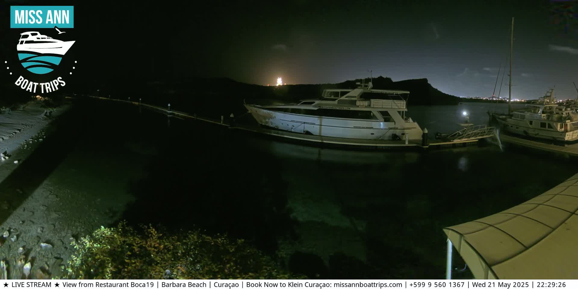 At night, several yachts are docked at a marina, with a hill visible in the background under a dark, clear sky.