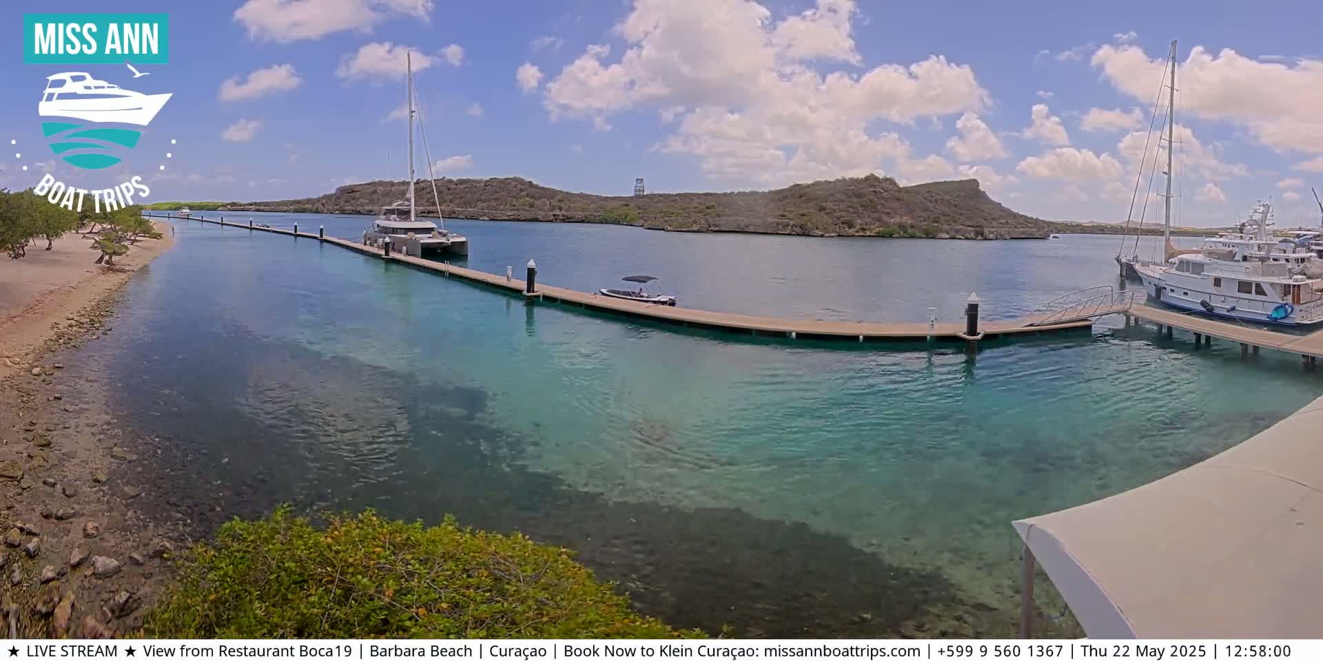 A marina with several boats docked under a partly cloudy sky shows clear turquoise water.
