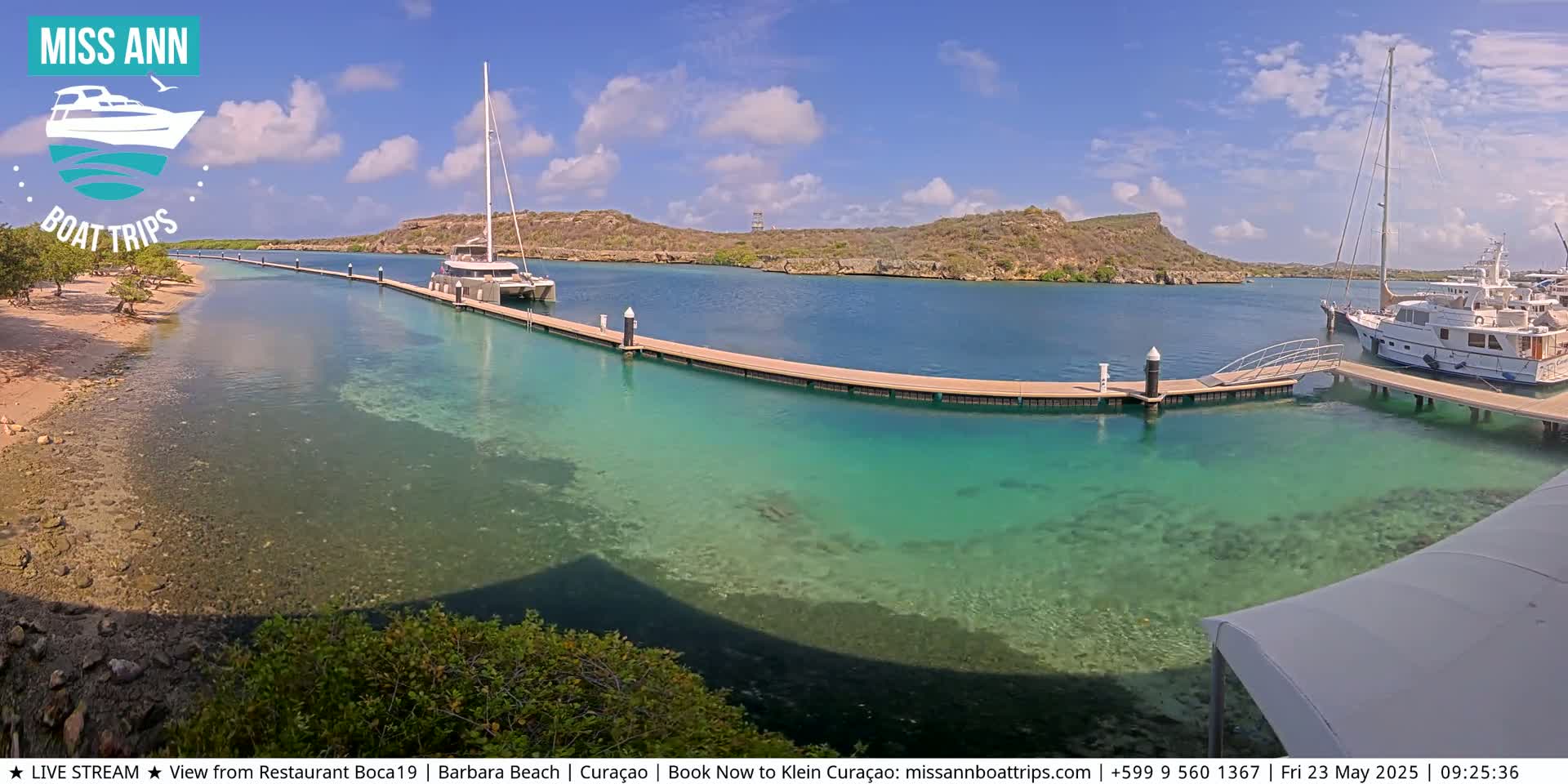 A sunny day shows a calm, clear turquoise body of water with several boats docked at a long pier, extending from a sandy shore lined with vegetation, and a hilly, dry landscape in the background.