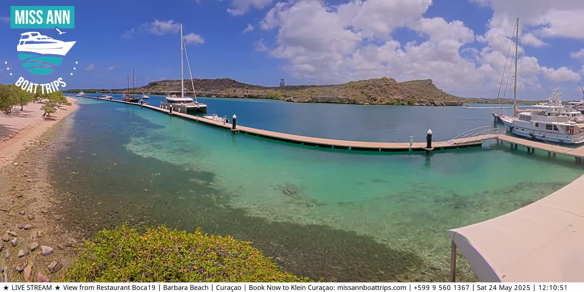 A sunny day shows a calm, clear turquoise bay with several boats docked at long piers, next to a sandy beach with sparse vegetation.