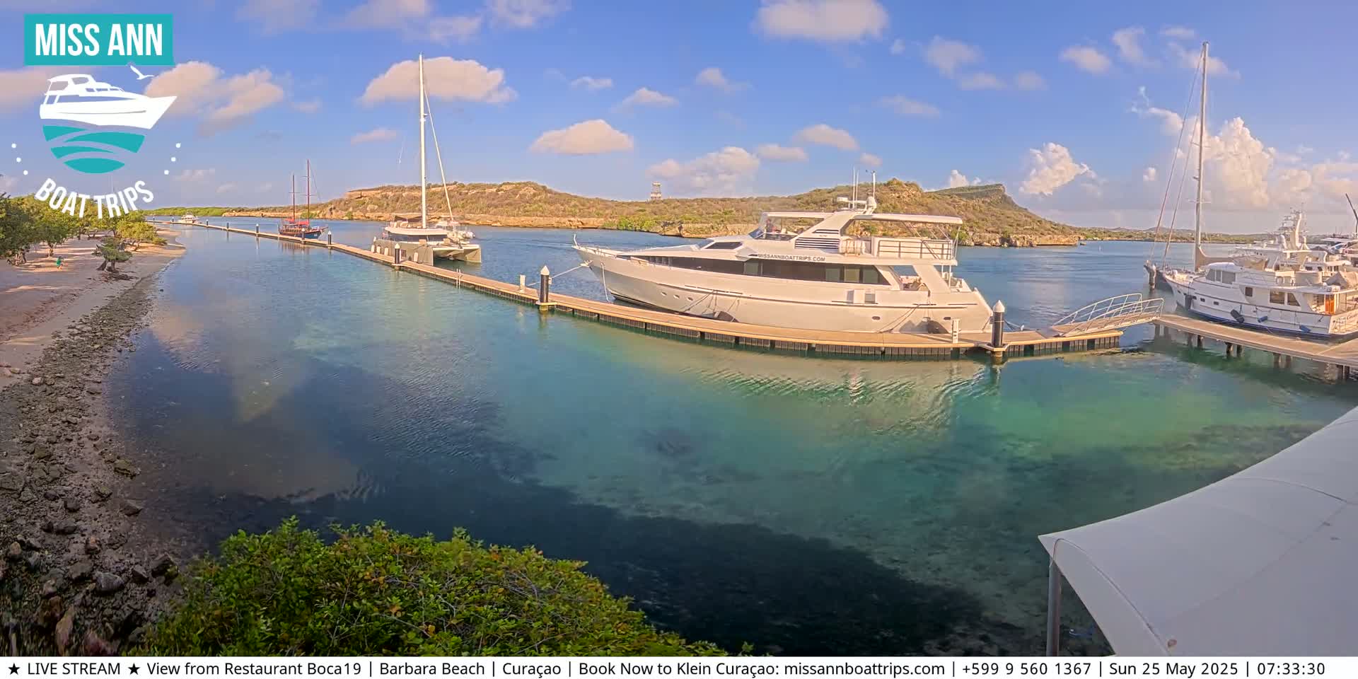 Several boats are docked at a marina on a sunny day with partly cloudy skies, the clear, shallow water showing the sandy bottom.