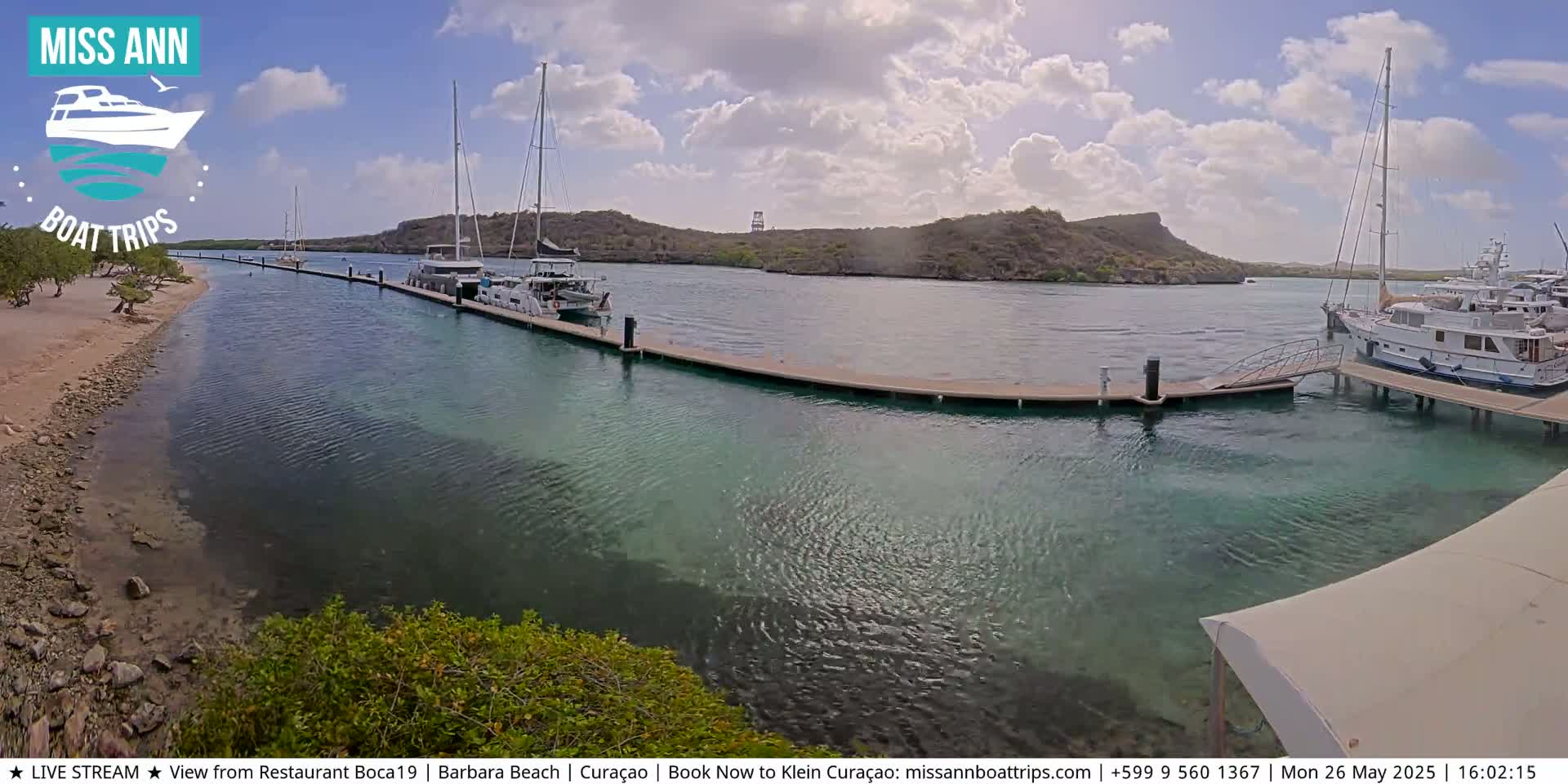 Several boats are docked at a marina on a sunny day with partly cloudy skies, next to a sandy shore.