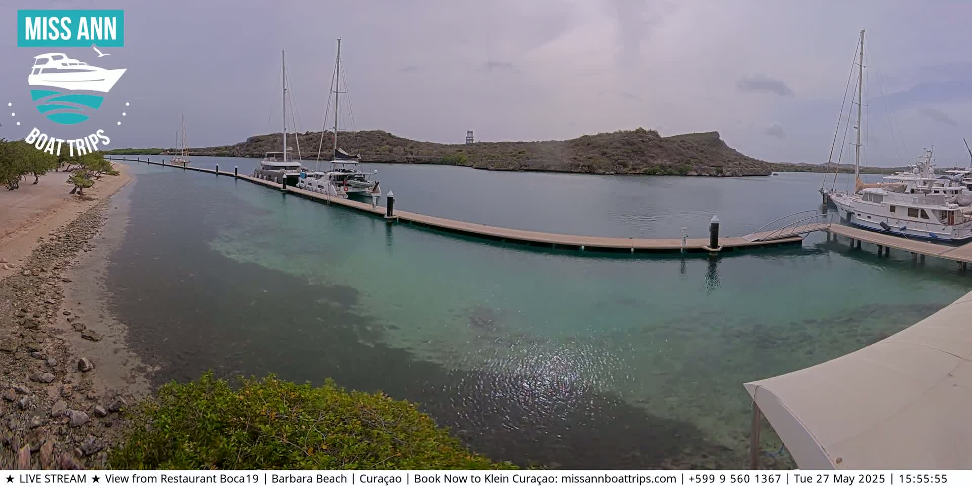 Several sailboats and motor yachts are docked at a marina on a partly cloudy day, with clear, shallow turquoise water visible around the docks and a sandy beach in the foreground.
