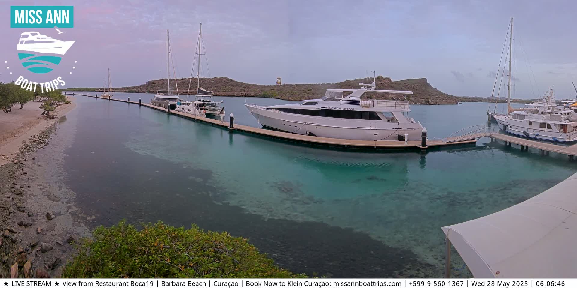 Several yachts and sailboats are docked at a marina with clear, shallow turquoise water, under an overcast sky.