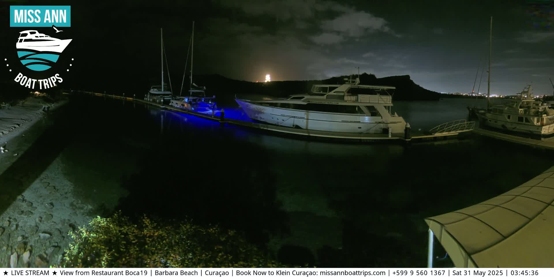 Several boats are moored at a marina at night under a dark, cloudy sky, with lights illuminating parts of the water and a distant shoreline visible.