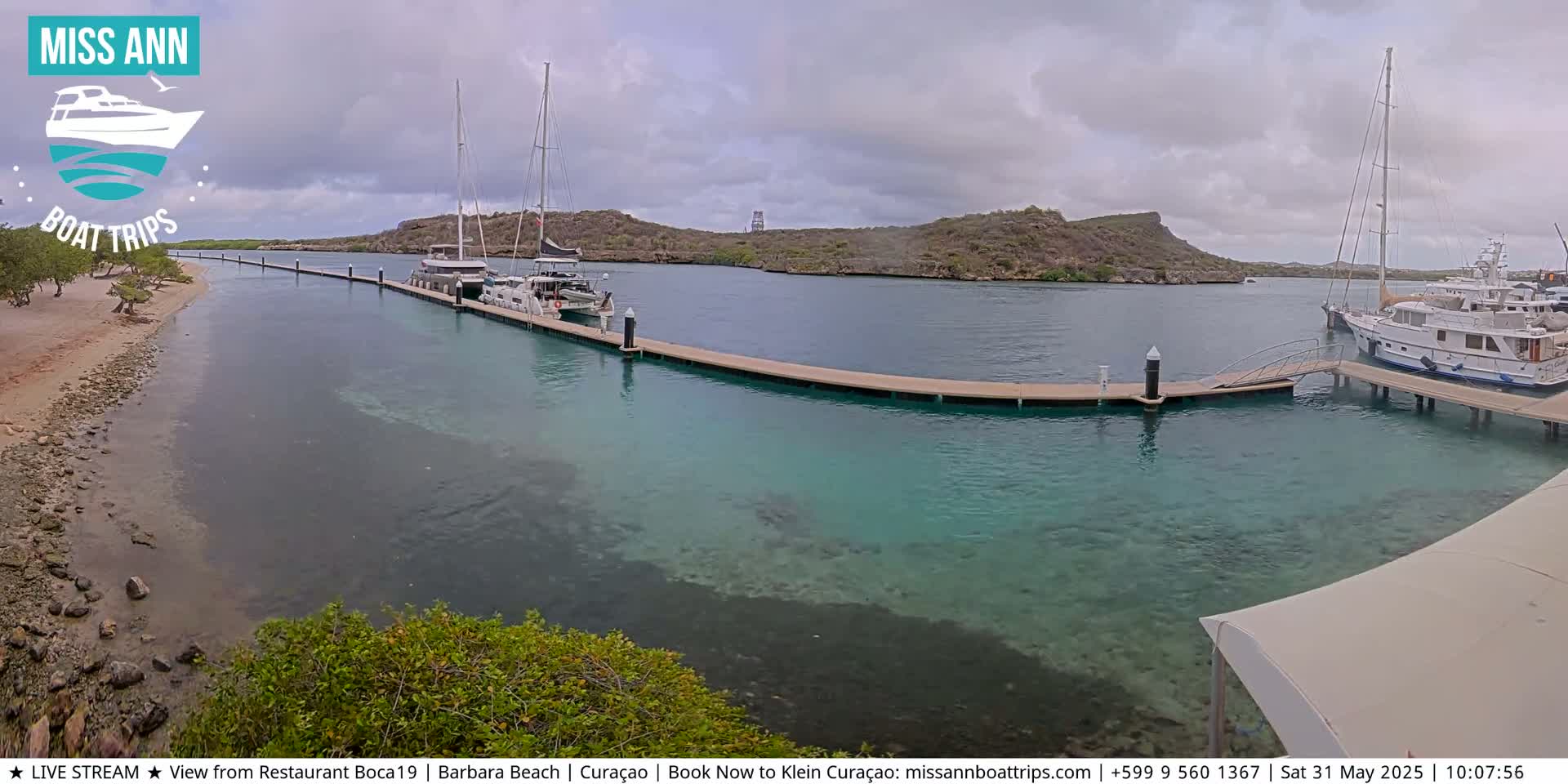 Several boats are docked at a marina with clear, shallow turquoise water, under a mostly cloudy sky.