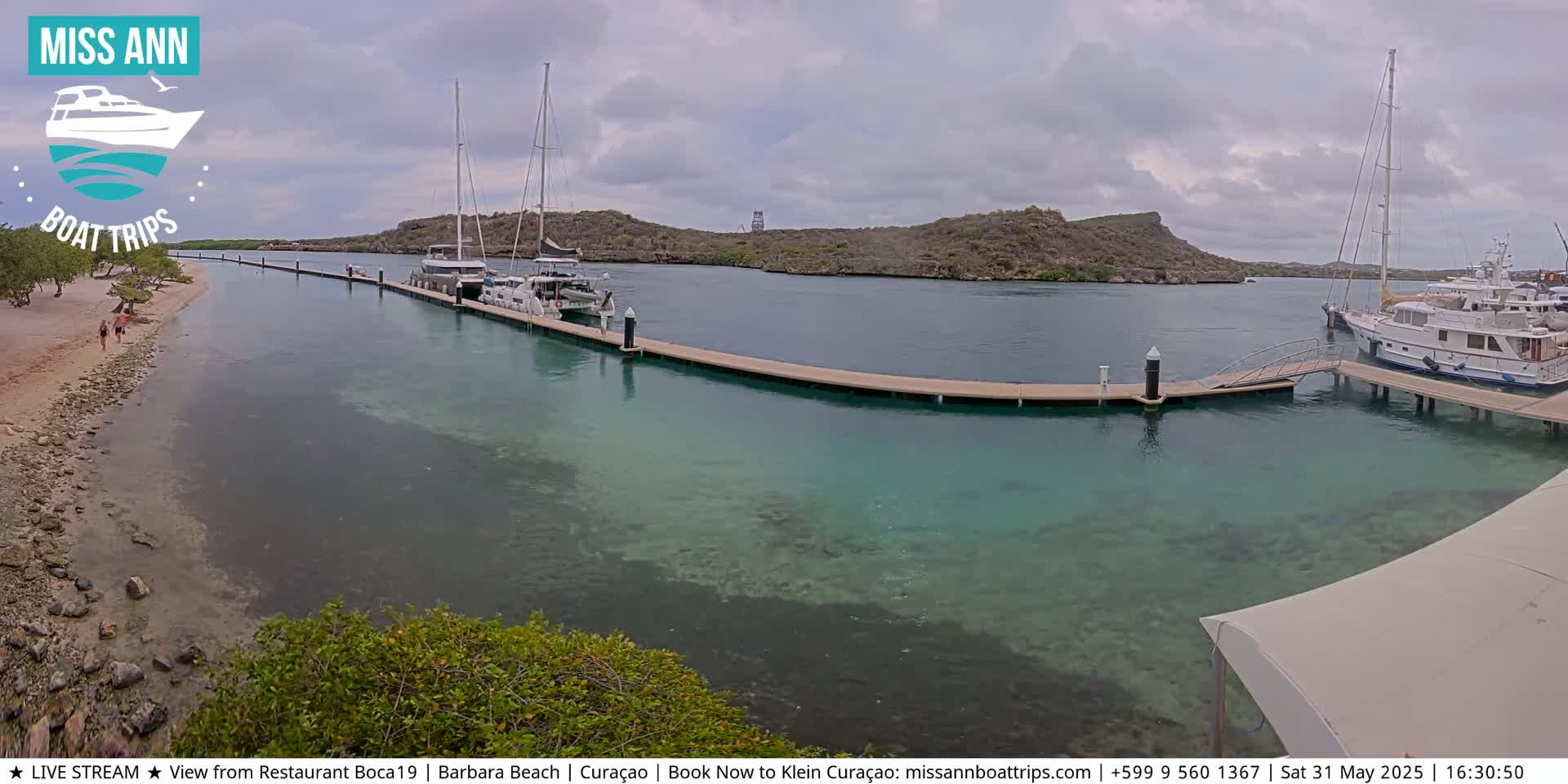 A calm, partly cloudy day reveals a marina with several boats docked along a long pier, a sandy beach with a few people, and a tranquil body of clear, teal water.