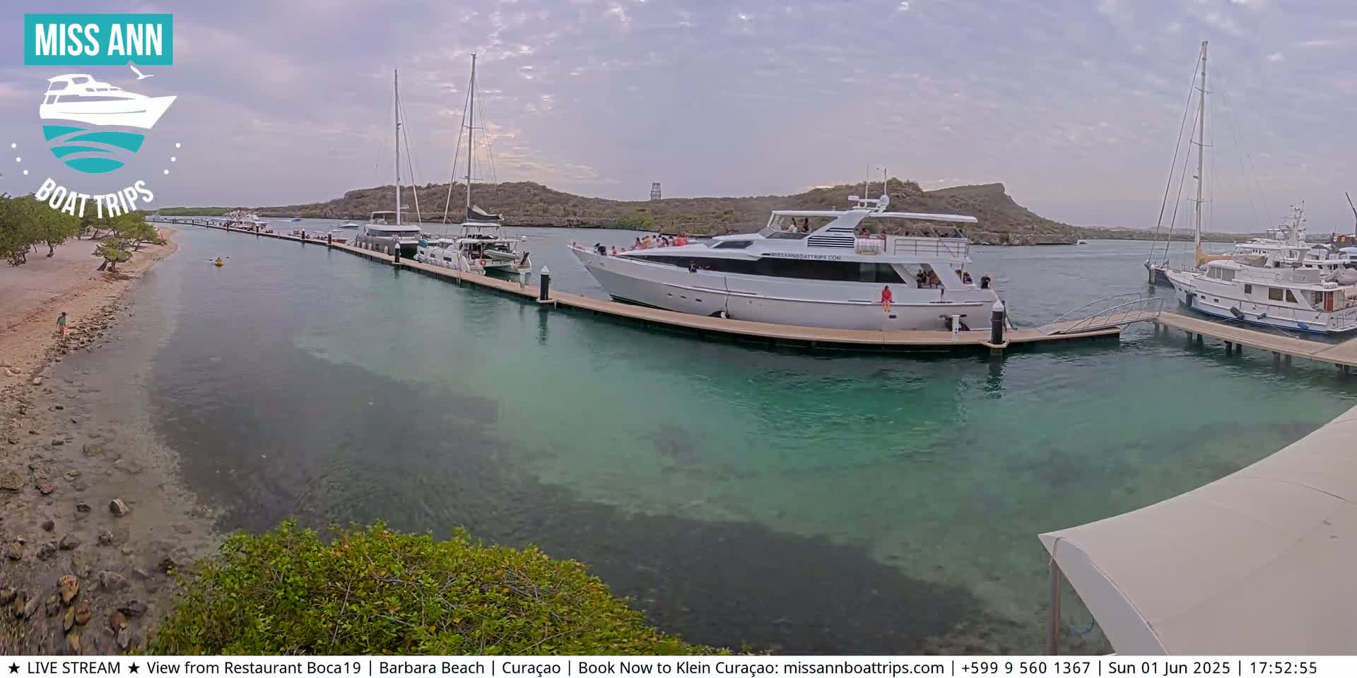 Several yachts and boats are docked at a marina on a partly cloudy day, with clear, shallow turquoise water visible.