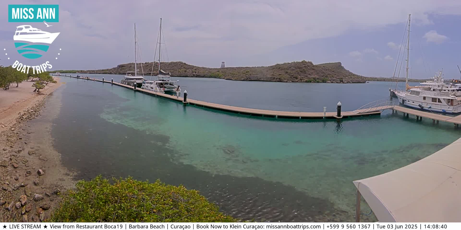 Several boats are docked at a marina with clear, shallow turquoise water, next to a sandy beach and arid hills under an overcast sky.