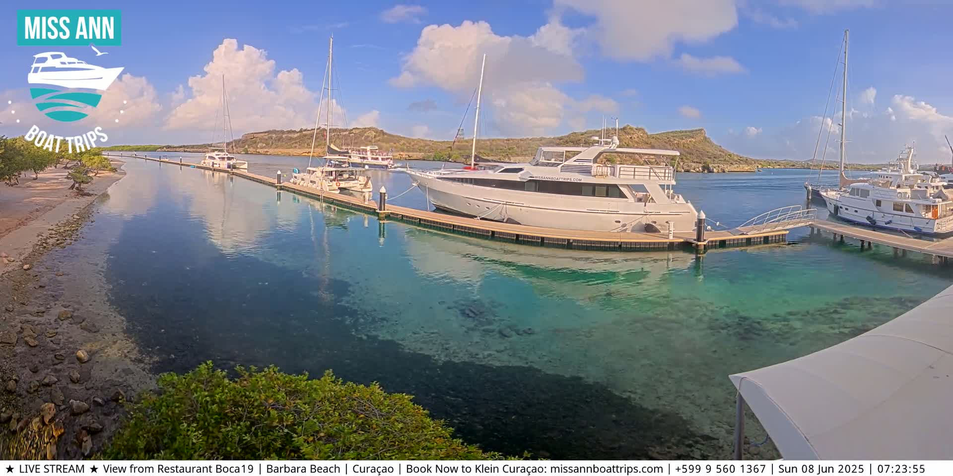 Several yachts and sailboats are docked in a marina with clear, shallow turquoise water under a partly cloudy blue sky.