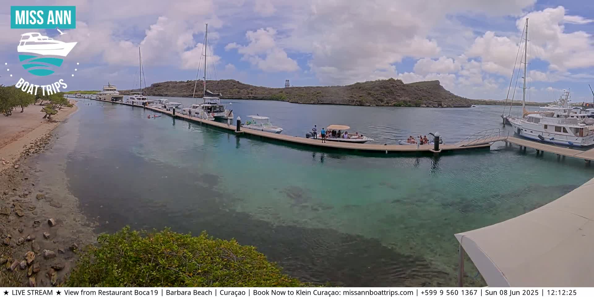 Several boats are moored at a dock in clear, shallow turquoise water near a sandy beach and rocky hills under a partly cloudy sky.