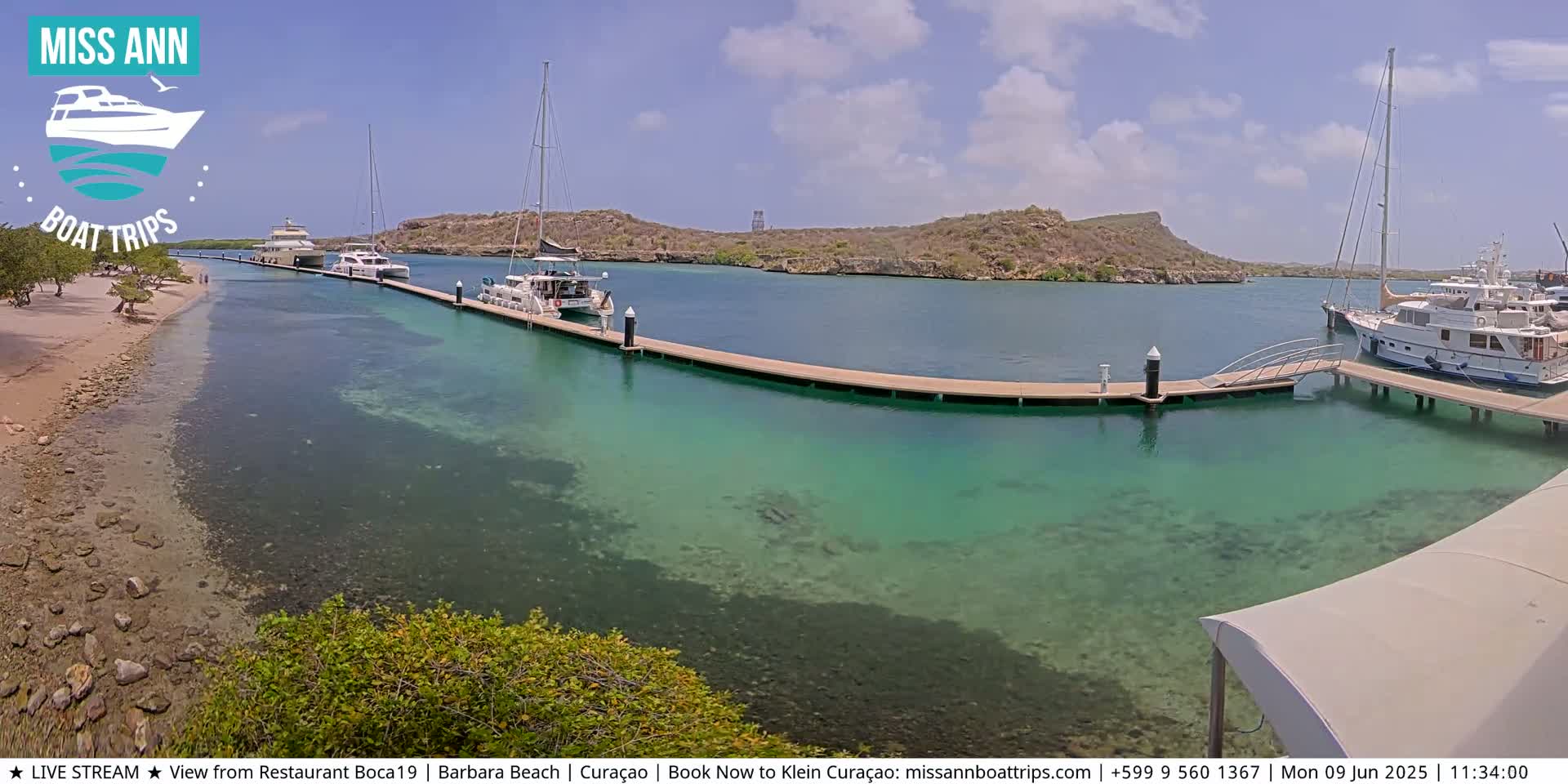 Several boats are docked at a marina with clear, shallow turquoise water, under a sunny sky.