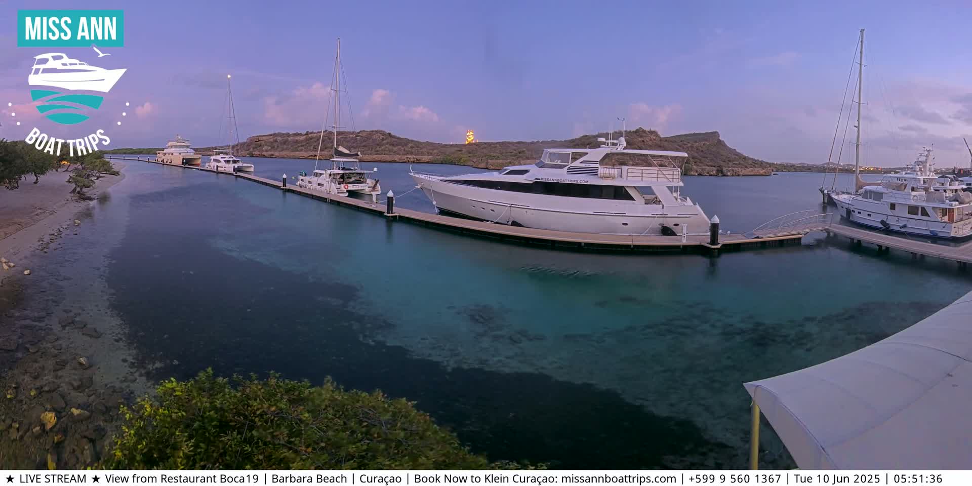 Several yachts and sailboats are docked at a marina at dusk, with calm, clear turquoise water surrounding them.