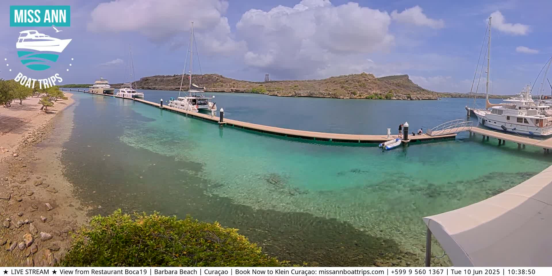 Several boats are docked at a marina with clear, shallow turquoise water, under a mostly sunny sky.