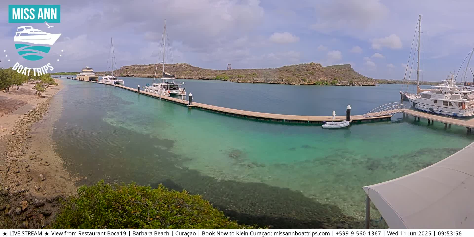 Several boats are docked at a marina with clear, shallow turquoise water, under a partly cloudy sky.