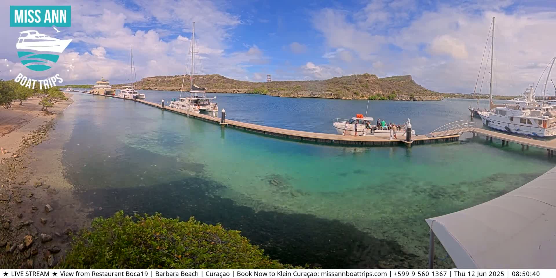 Several boats are moored at docks in a calm, clear turquoise harbor on a sunny day, with hilly terrain visible in the background.