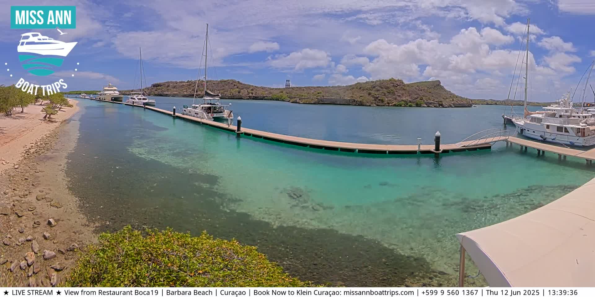 A sunny day shows a tranquil marina with several boats docked at a pier, clear turquoise water, and a sandy beach in the background.