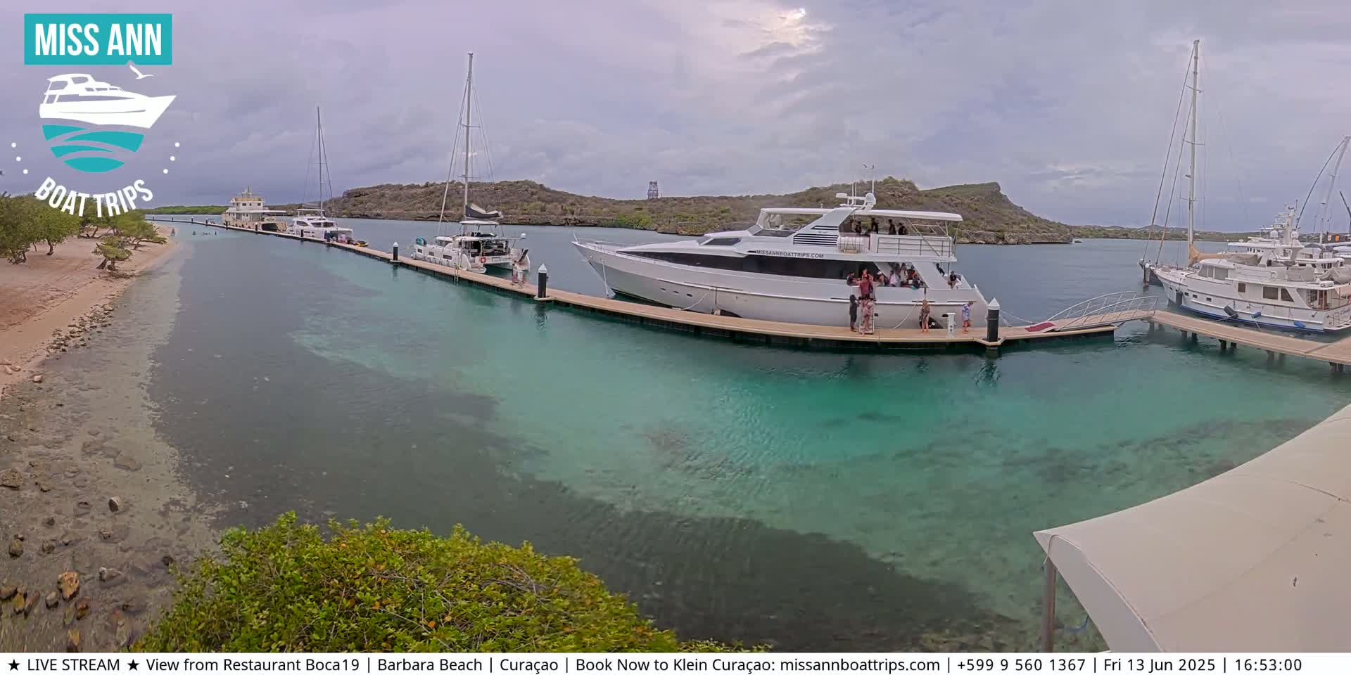 Several boats are docked at a marina on a partly cloudy day, with clear, shallow turquoise water visible.