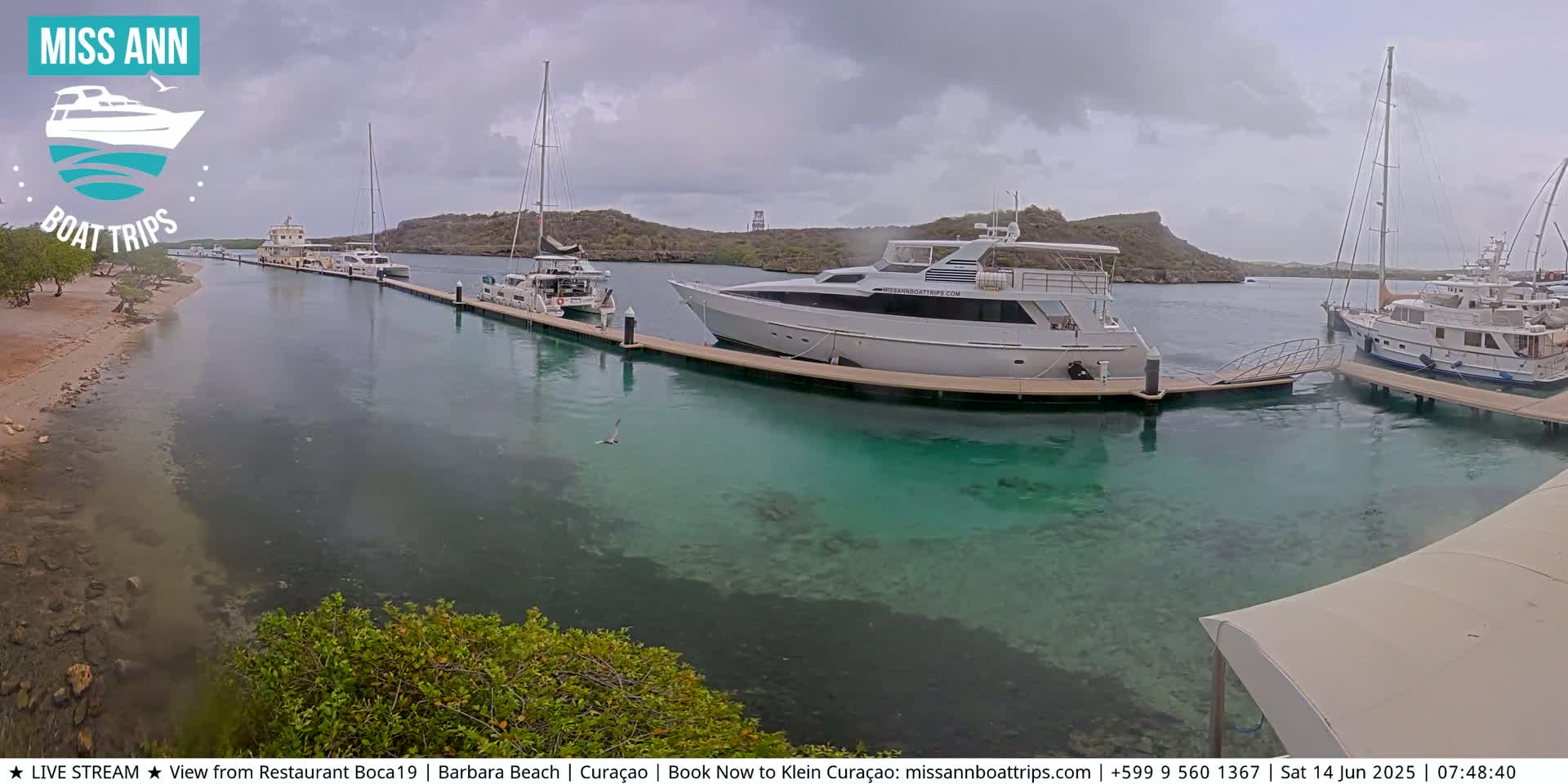 Several boats are docked in a calm, clear, turquoise harbor under an overcast sky.